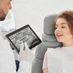 Na zdjęciu dentysta w Michałowicach prezentuje pacjentce zdjęcie rentgenowskie jej zębów przed przeprowadzeniem zabiegu wstawienia implantu tytanowego. To krok w procesie planowania skutecznej terapii implantologicznej.